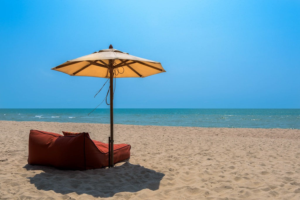 Large beach umbrella shading low seats beside bright sand and blue water