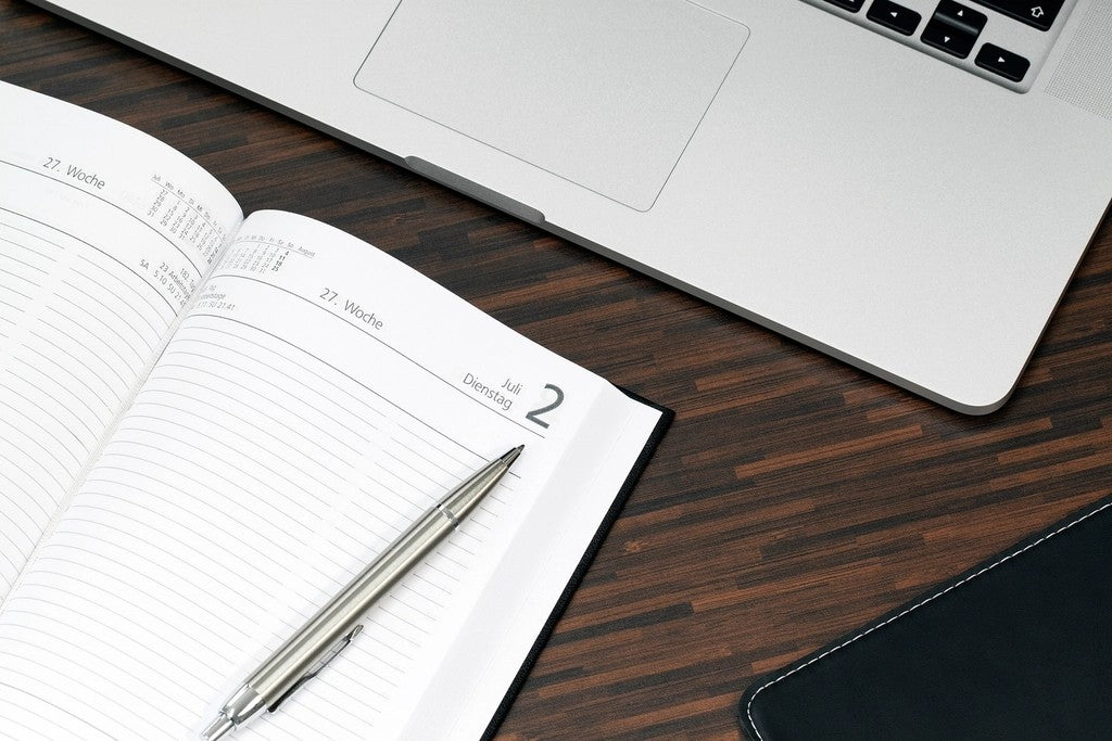 Blank weekly paper planner with a pen beside a silver laptop on a dark wood desk