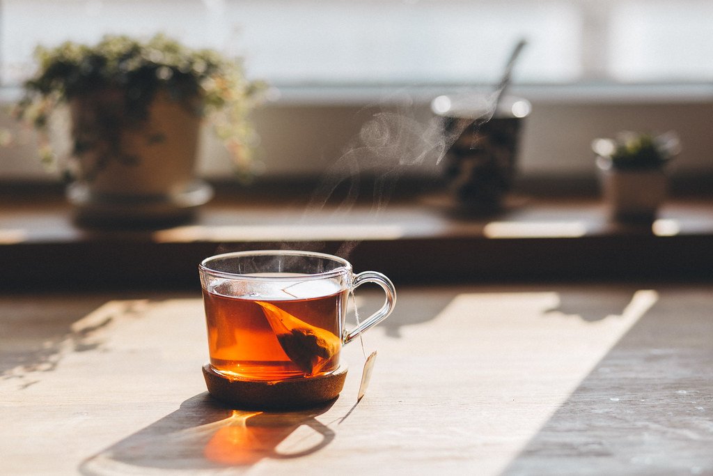 A clear glass mug of warm tea on a cork coaster on a sunlit wooden counter