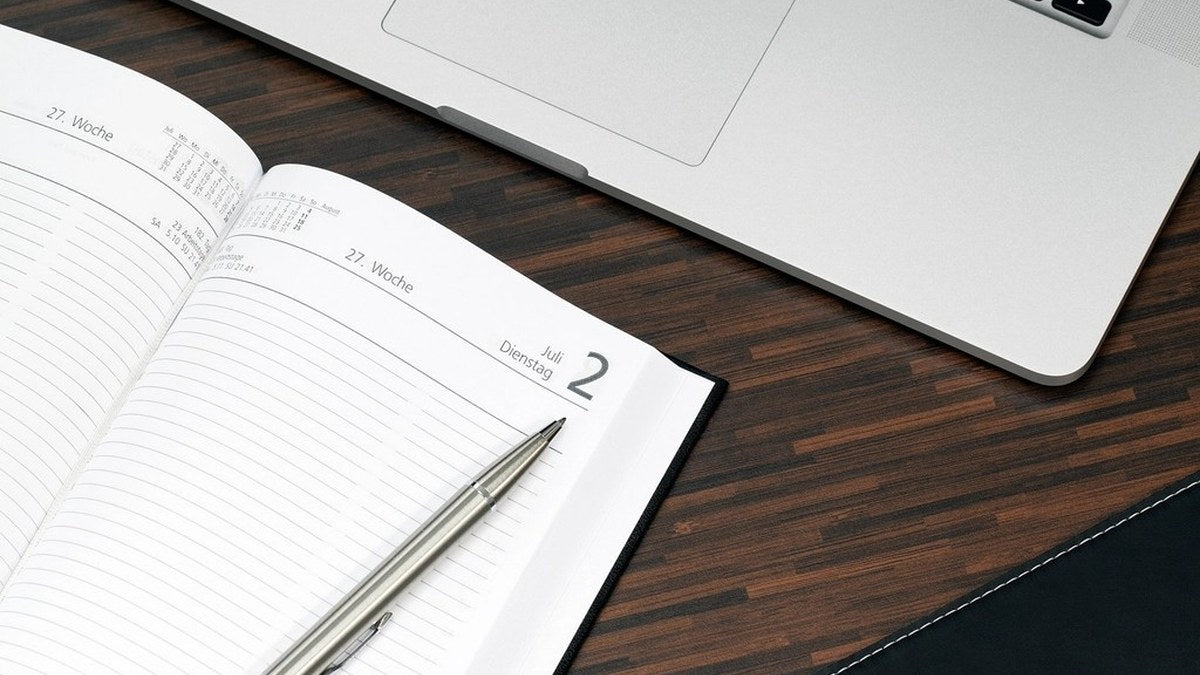 Weekly paper planner with a pen beside a laptop on a dark wood desk