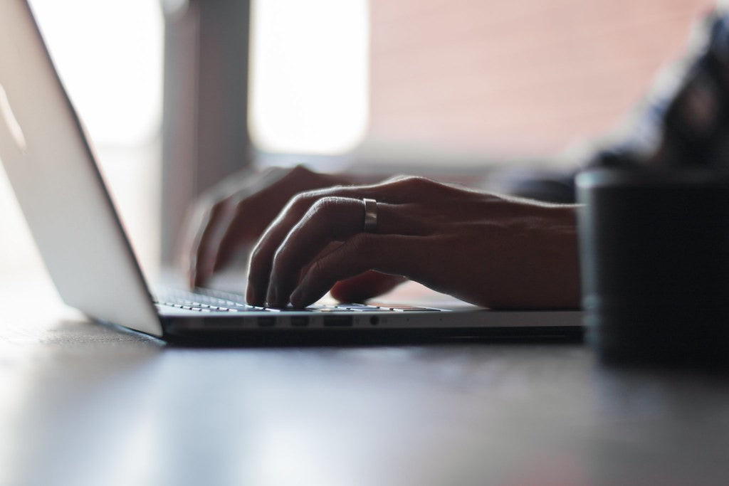Close-up of a person typing on a laptop at a table beside a dark mug with soft daylight behind them