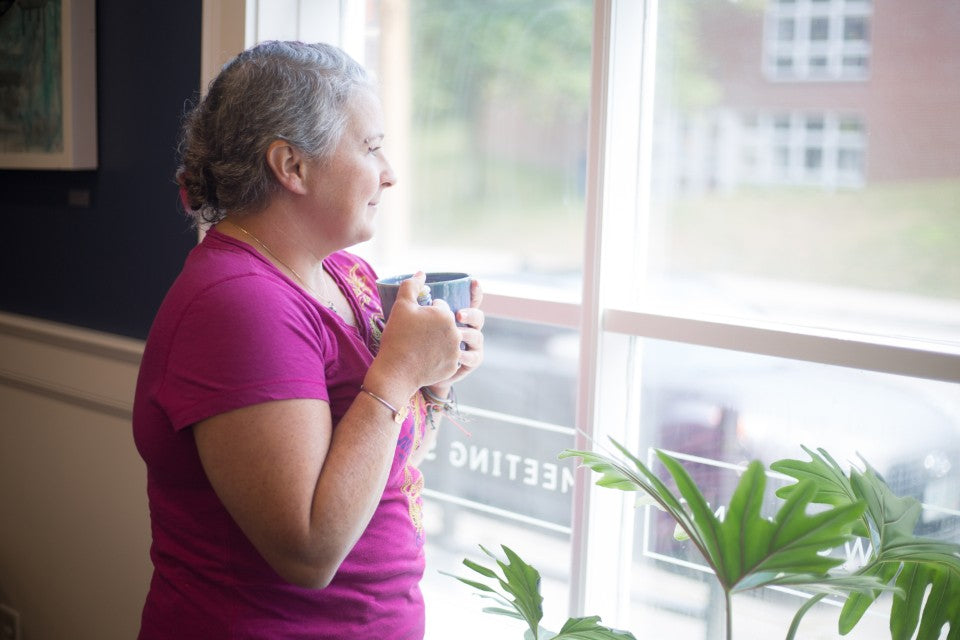 Woman by a bright window holding a mug with a small craft in hand