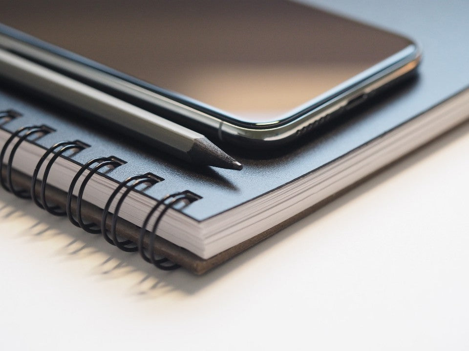 Smartphone resting on a dark spiral notebook beside a sharpened pencil