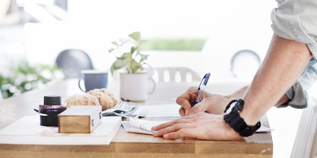 Person writing on a small set of papers at a bright wooden table