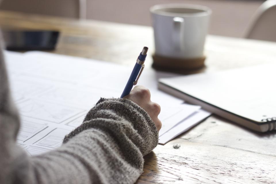 A person writing notes with a pen in a notebook on a wooden desk next to a warm mug