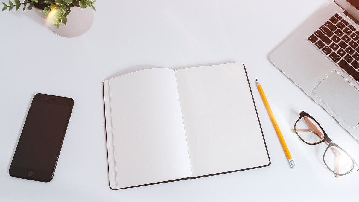 Open notebook, phone, pencil, glasses, and laptop arranged on a bright work desk